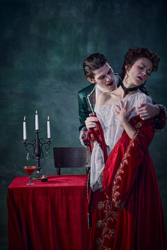 Portrait of young man in image of medieval prince, vampire biting innocent young woman over dark green background. Blood drinking