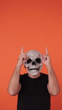 Person with skull and crossbones mask celebrating Halloween, pointing up with fingers, on orange background. Celebration concept, All Souls' Day and All Saints' Day.