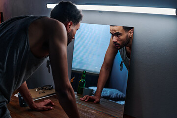 Young man with post traumatic disorder bending over table and looking at his reflection in mirror...