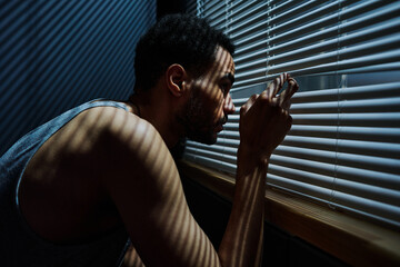Side view view of young sleepless man looking through venetian blinds on window during sneak peek...