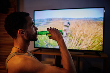 Young man with post traumatic disorder having beer while watching video about military operation...