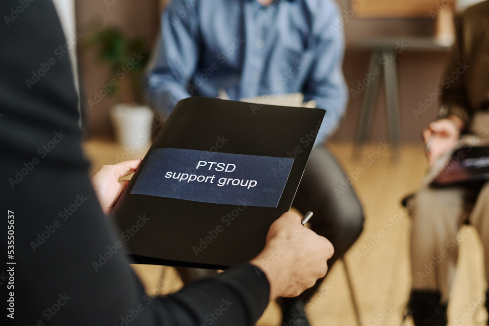 Wall mural Selective focus on black folder with sticker saying PTSD support group held by young male patient of psychotherapist sitting in front