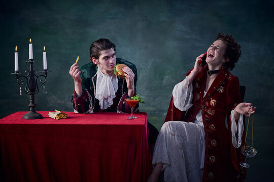 Portrait of man and woman in image of medieval vampires over dark green background. Man eating burger, woman talking on phone