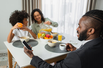 Fototapeta na wymiar african american man in suit using smartphone with blank screen during breakfast with family on blurred background.