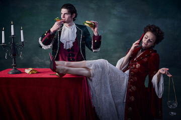 Portrait of man and woman in image of medieval vampires over dark green background. Man eating burger and drinking bloody mary, woman talking on phone