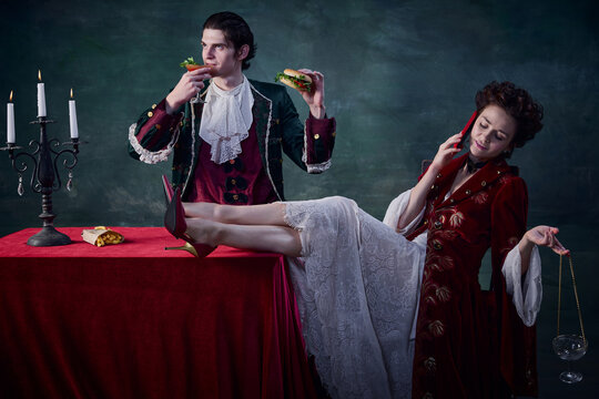 Portrait of man and woman in image of medieval vampires over dark green background. Man eating burger and drinking bloody mary, woman talking on phone
