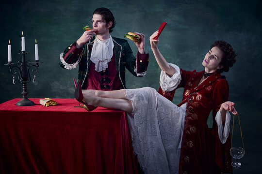 Portrait of man and woman in image of medieval vampires over dark green background. Man eating burger, woman drinking and taking selfie