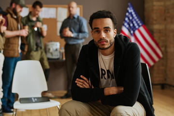 Young unhappy man with post traumatic syndrome looking at camera while sitting against group of...