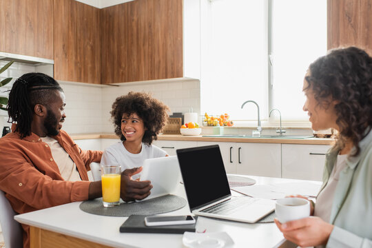 happy african american woman holding cup near laptop while husband and daughter talking near digital tablet in kitchen.