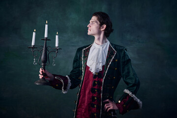 Portrait of man in noble medieval costume in image of vampire holding candles isolated over dark green background. Hunter