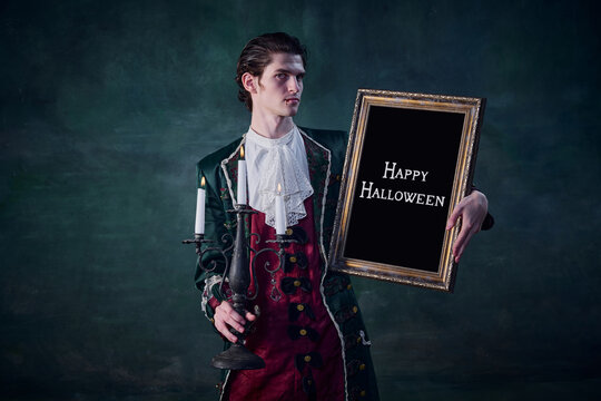 Portrait of man in noble medieval costume in image of vampire holding picture frame isolated over dark green background. Hunter