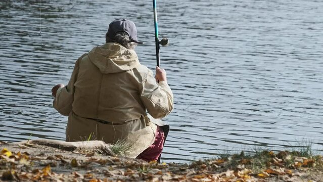 The fisherman catches fish on the river bank. Back view of a lone elderly fisherman sitting with a fishing rod on a sunny autumn day. Calm and peaceful picture. Lifestyle, leisure activity on nature