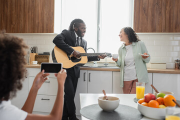 curly african american kid taking photo of father in suit playing acoustic guitar near happy wife in kitchen.
