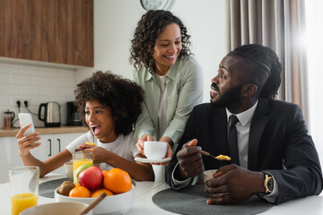cheerful african american woman holding cup of coffee near husband in suit while preteen daughter using smartphone.
