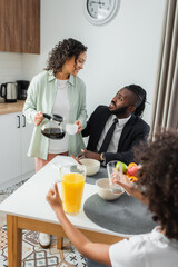 Fototapeta na wymiar happy african american woman holding coffee pot and cup while looking at husband in suit near preteen daughter during breakfast.