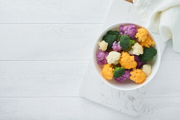 Colorfu cauliflower. Cauliflower cut into small pieces white bowl on light stone marble concrete background. Purple, yellow, white and green color cabbages. Agricultural harvest. Mock up. Top view.