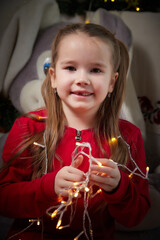 Portrait of a little pretty girl with garland and long beautiful hair in red dress in the room decorated for Christmas. Christmas and New Year holiday