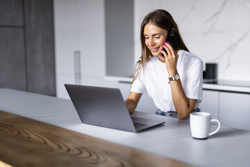 Photo of a young cheerful happy woman at home at the kitchen using laptop computer talking by mobile phone.