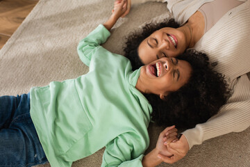 top view of happy african american girl laughing while lying on carpet with cheerful mother.