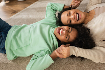positive african american girl laughing while lying on carpet with cheerful mother.