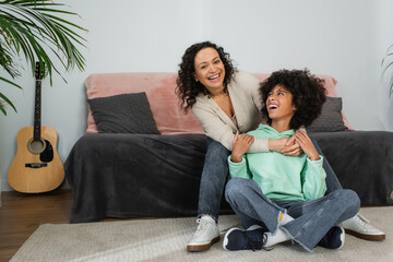 curly african american mother smiling while hugging excited preteen daughter sitting with crossed...