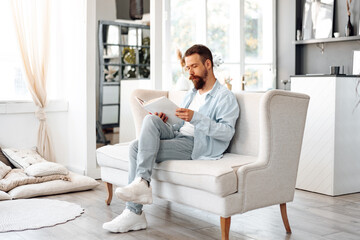 Young bearded handsome man reading book on couch at home