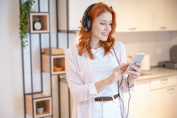 Young, red hair, pretty girl in good mood and bright outfit enjoying listening to the music with headphones on her head, while using her smartphone and standing at home