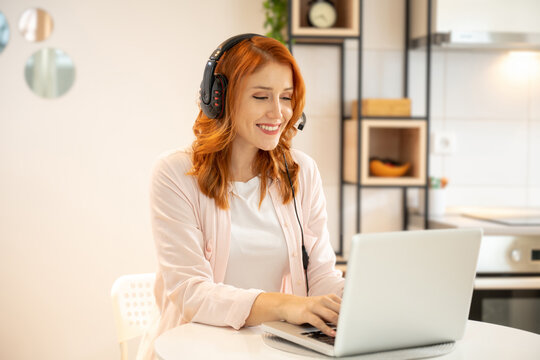 Young Smiling Friendly Ginger Girl Working As Agent At Call Center From Home. Wearing Headset And Typing On Laptop.