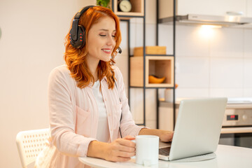 Fototapeta na wymiar Young smiling friendly ginger girl working as agent at call center from home while drinking coffee. Wearing headset and typing on laptop.