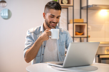Young smiling man working on laptop at home. Wearing casual clothes and drinking coffee.