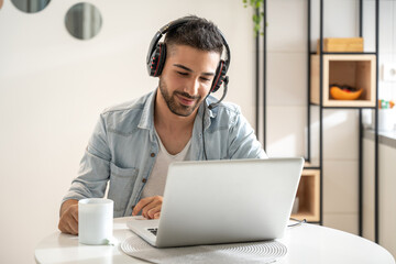 Young friendly man working as agent at call center from home while drinking coffee. Wearing headset...