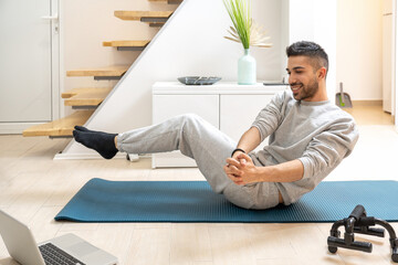 Fototapeta na wymiar Happy young handsome man doing Russian Twist exercise on exercise mat and watching sport classes via laptop at home.