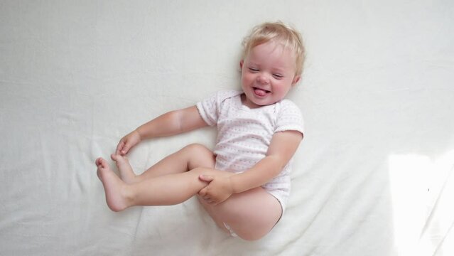 Upper view of charming funny cute baby in diaper and bodysuit lying on back playing with feet, child having fun laughing at camera in bed on white sheet.