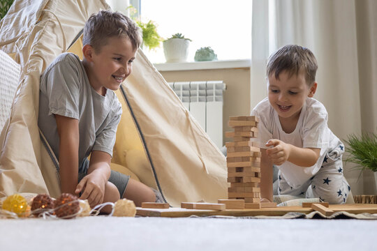 Two male children playing Jenga hardwood bricks tower construction childish room with linen wigwam