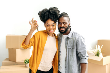 Happy married african american couple, newlyweds, bought their first own home, stand in living room against the background of cardboard boxes with things, look at camera, smile,the girl shows the keys