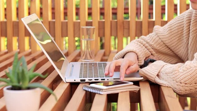Side view of faceless anonymous woman freelancer wearing beige jumper and glasses working on notebook and using mobile phone, checking e-mail, swiping on cell phone.