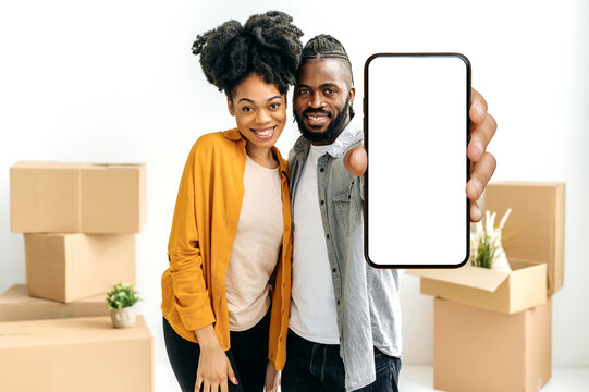 Happy Married African American Couple, Newlyweds, Show Smart Phone With Empty White Mock-up Screen While Stand In Living Room Against The Background Of Cardboard Boxes With Things In Their New Home