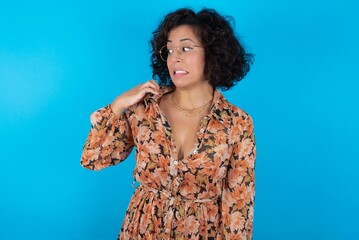 young brunette woman with curly hair wearing flowered dress standing over blue background stressed, anxious, tired and frustrated, pulling shirt neck, looking frustrated with problem