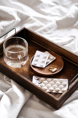 Wooden tray with pills, vitamins or supplements and water glass on the bed. Health care in the morning at home.