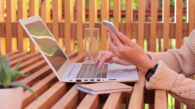 Anonymous faceless woman wearing beige jumper sitting at her workplace, typing on laptop and using smart phone for working, faceless female freelancer with notebook and mobile phone.