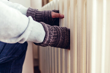 Cold home, freezing. Using heater at home in winter. Senior man warming his hands sitting by device and wearing warm clothes. Heating season. Man warms up hands over heater.