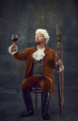 Tasting red wine. Vintage portrait of young man in brown retro suit and white wig like medieval...