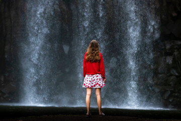 A girl in a red sweatshirt and red and white skirt stands over a powerful tropical waterfall in australia; millaa millaa falls in queensland; queensland rainforest