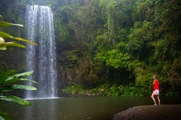 A girl in a red sweatshirt and red and white skirt stands over a powerful tropical waterfall in...