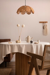 Minimalist composition of dining interior with round wooden table, design chair, pendant rattan lamp, vase with dried flowers, decoration and personal accessories. Stylish home decor. Template.