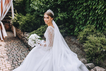 Fototapeta premium A beautiful bride in a long white dress with a bouquet in her hands, a diadem on her head, sits in the park, against the background of a wall with green ivy. Close-up wedding photography, portrait.