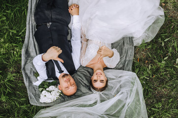 A stylish groom and a beautiful bride with a diadem lie on a plaid with a long white veil, a...