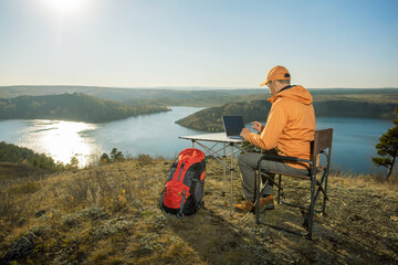 Fototapeta premium Man working on laptop on top mountains and relaxes near river at sunset. Freedom, remote work, freelancer, technology, internet, travel and vacation concepts.