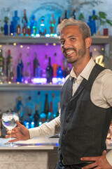 Attractive young barman in a white shirt and black apron smiling looking to camera at party in nightclub. Night lifestyle concept.