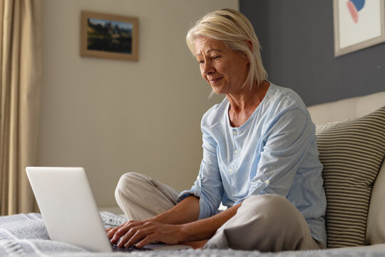 Happy senior caucasian woman sitting on bed in bedroom, using laptop